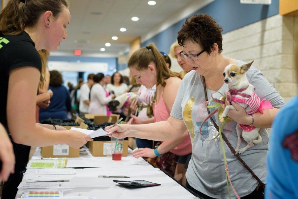 people in lined up in front of a table registering for a event woman in forefront holding a fawn and whit chihuahua in a pink dress