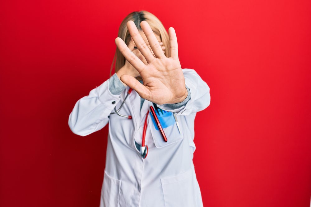 a woman in a white coat and a stethoscope around her neck holding hand in front of her face and one out to block the camera on a red background